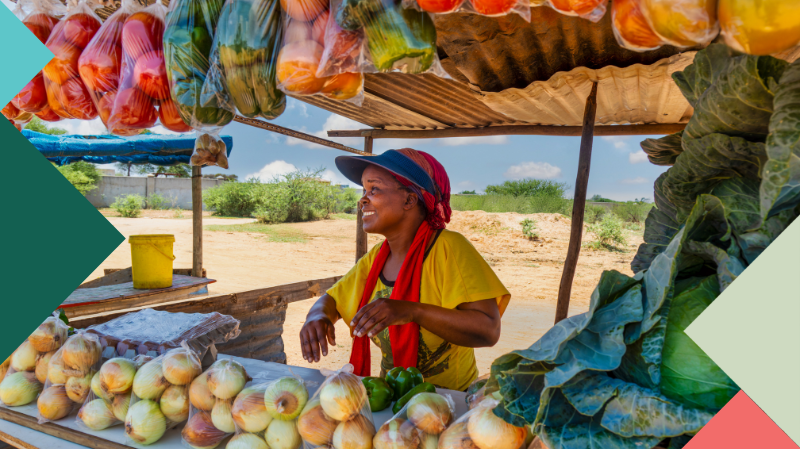 A woman in Africa stands behind a stall full of fruit and vegetables smiling and selling her stock.